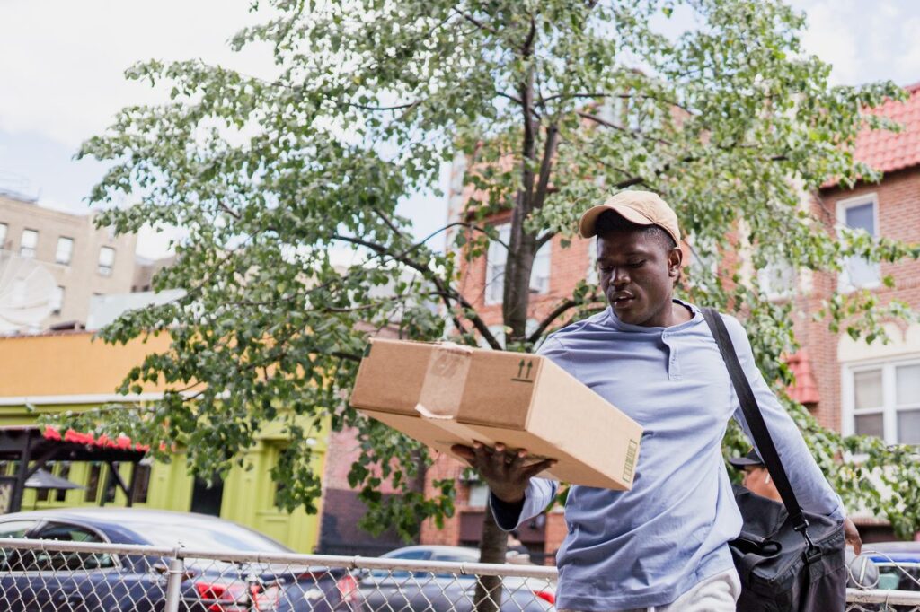 A delivery man walks through an urban neighborhood carrying a cardboard package amidst residential buildings.